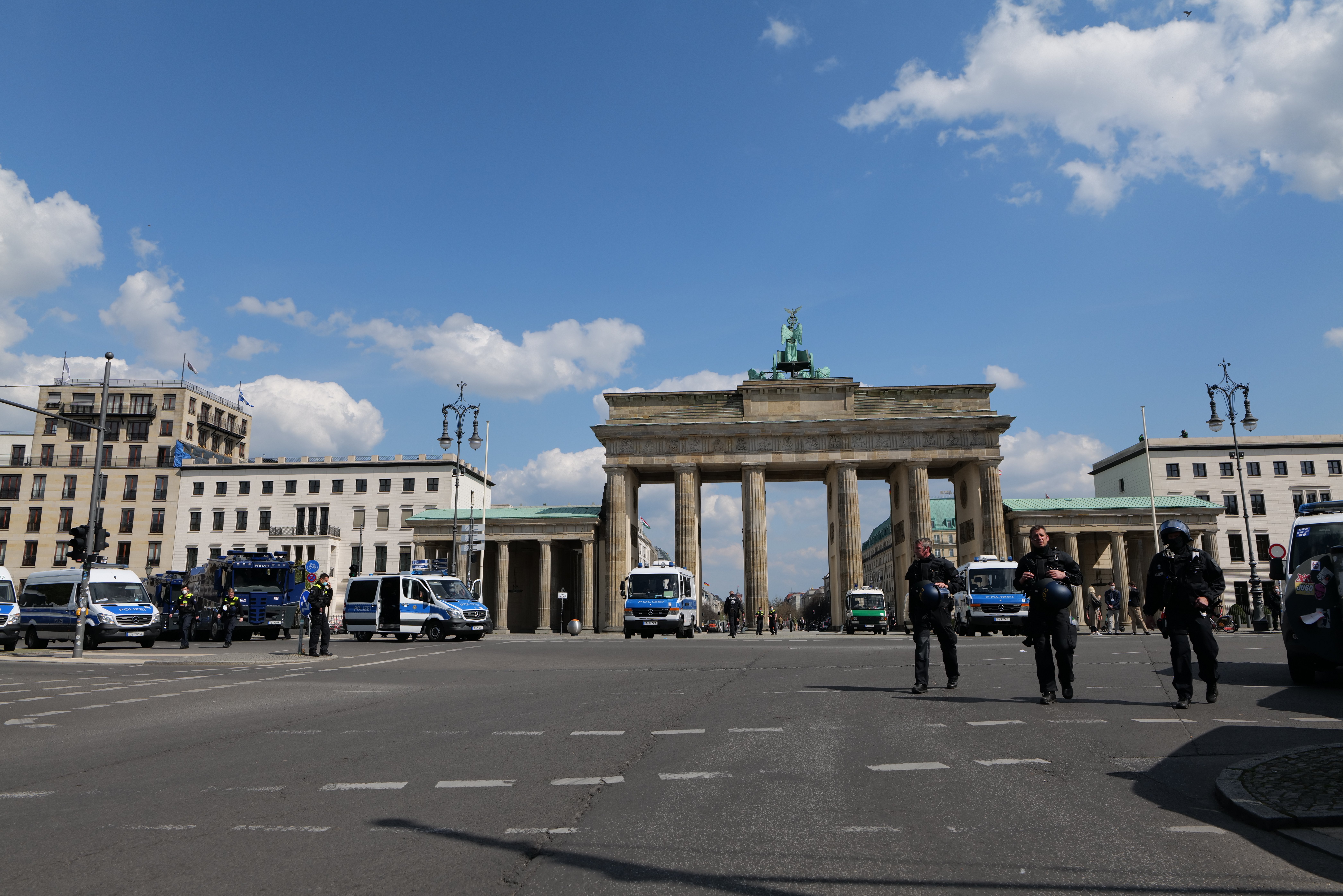 Eine Gruppe von Polizisten vor dem Brandenburger Tor in Berlin, Deutschland, das mit Säulen, einer Statue und umgeben von Gebäuden mit Fenstern, Fahrzeugen auf der Straße, Laternenmasten und Ampelstangen mit Masten unter einem bewölkten Himmel.