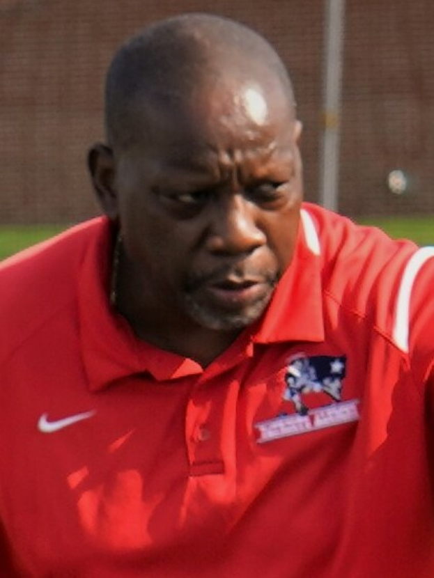 Ein Mann in einem roten T-Shirt steht auf einem Feld und hat die Hände in die Hüften gestützt, er sieht ernst aus, der Hintergrund ist unscharf.