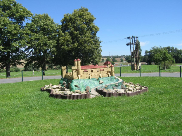 Minigolfplatz mit Rasenfeld, eingezäunt mit Pfählen und Ketten, Bäumen, Strommasten mit Drähten und einem bewölkten Himmel im Hintergrund.