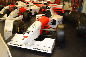 Eine Ausstellung von Rennwagen in einem Museum mit Menschen, die zuschauen, mit einem Textschild, einer Seilabsperrung und einer Hintergrundwand.