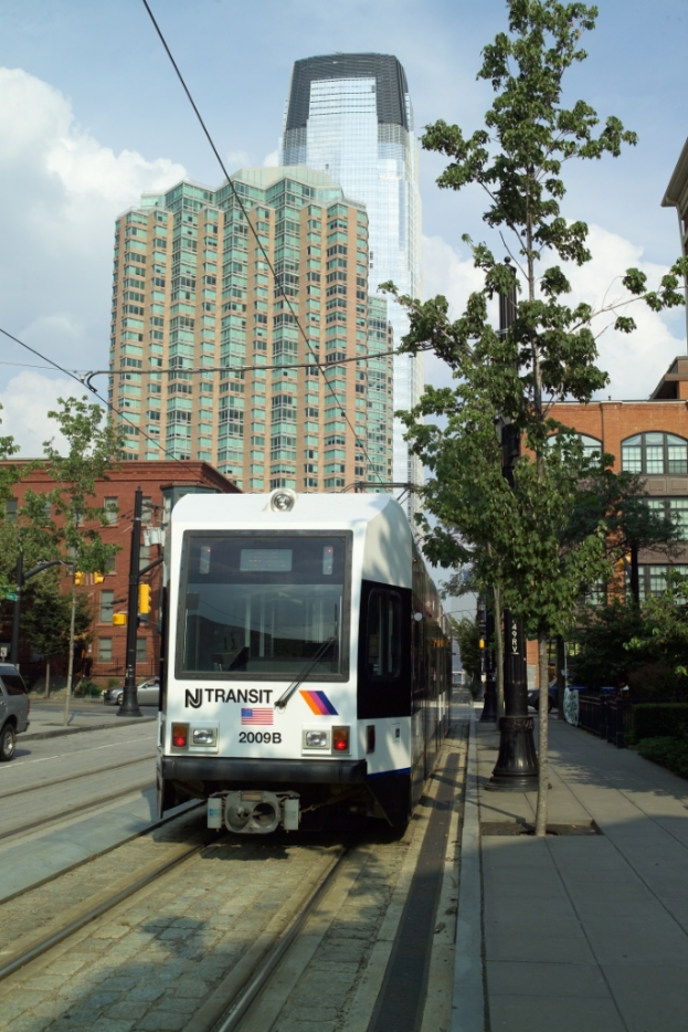 Ein Zug auf Eisenbahnschienen mit Oberleitungen, flankiert von B├Ąumen, Masten und Geb├Ąuden, mit einem Wolkenkratzer und Himmel im Hintergrund.