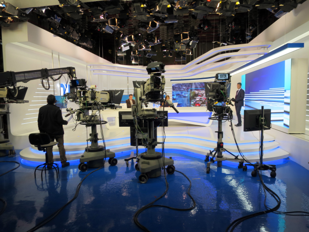 Ein TV-Studio mit mehreren Kameras auf dem Boden, einer sitzenden Person im Vordergrund und einigen stehenden Personen im Hintergrund, mit an der Decke montierten Scheinwerfern für eine Livesendung.