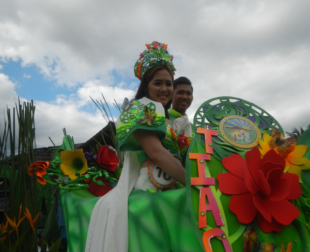 Ein Mann und eine Frau auf einem grünen und weißen Paradewagen, der mit Blumen und Text geschmückt ist, wobei die Frau ein grünes Kleid trägt und der Mann hinter ihr steht, vor einem Hintergrund aus Bäumen und einem klaren blauen Himmel.