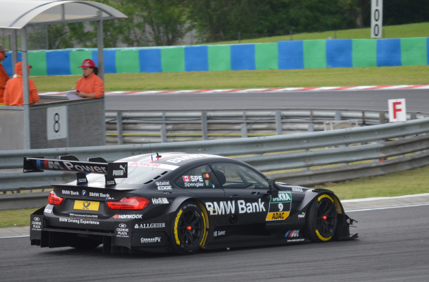 Ein BMW M4 DTM-Rennwagen f├╝hrt auf einer Rennstrecke, umgeben von einem Zaun, Gras, einem Schild, einem Schuppen und einigen Menschen in orangefarbenen Jacken und M├╝tzen, mit B├Ąumen im Hintergrund.