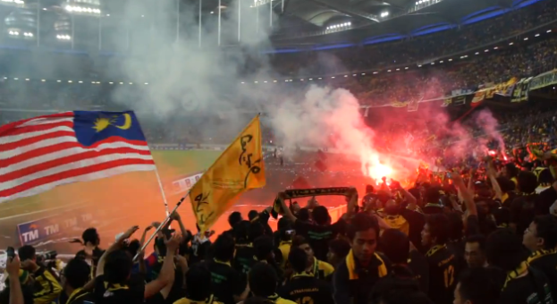 Eine große Menge Menschen in einem Stadion mit Bengalos, Fahnen und einer Kamera, mit Rauch und Schildern, die "Malaysia vs Malaysia Live Streaming" lesen.