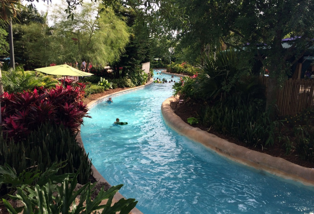 Ein Schwimmbecken mit schwimmenden Menschen, umgeben von Pflanzen, Bäumen und einem Zaun, mit Menschen, Sonnenschirmen und Gebäuden im Hintergrund.