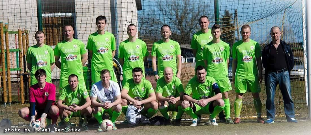 Gruppe von Männern in grünen T-Shirts, die auf einem Fußballfeld stehen oder knien, mit einem Ball und einem Torpfosten umgeben von Bäumen, Gebäuden und Fahrzeugen.