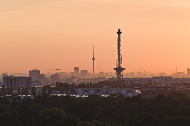 Ein Blick auf die Berliner Skyline bei Sonnenuntergang von einem Hügel aus, mit Gebäuden, Bäumen und dem Fernsehturm im Hintergrund, mit einem Himmel voller Oranges, Pinks und Purpurs.