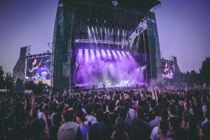 Eine große Menge Menschen bei einem Musikfestival schaut auf eine beleuchtete Bühne mit Instrumenten und Bildschirmen, die vor einem Gebäude, Bäumen und einem klaren blauen Himmel aufgebaut sind.