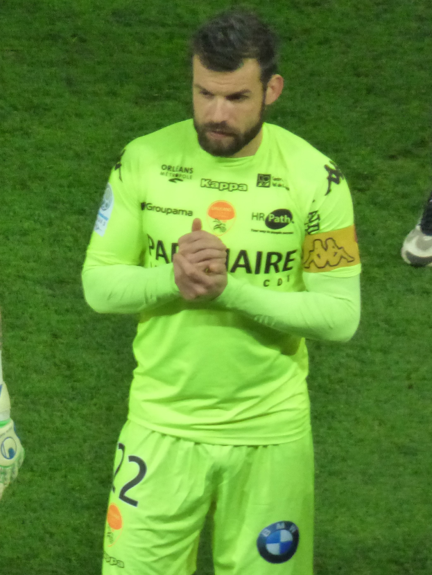 Ein Mann in einem gelben Torwarttrikot, der eine grüne Shorts und ein T-Shirt trägt, steht auf einem Fußballfeld und klatscht in die Hände, wobei auf der linken Seite des Bildes das Bein einer anderen Person zu sehen ist.