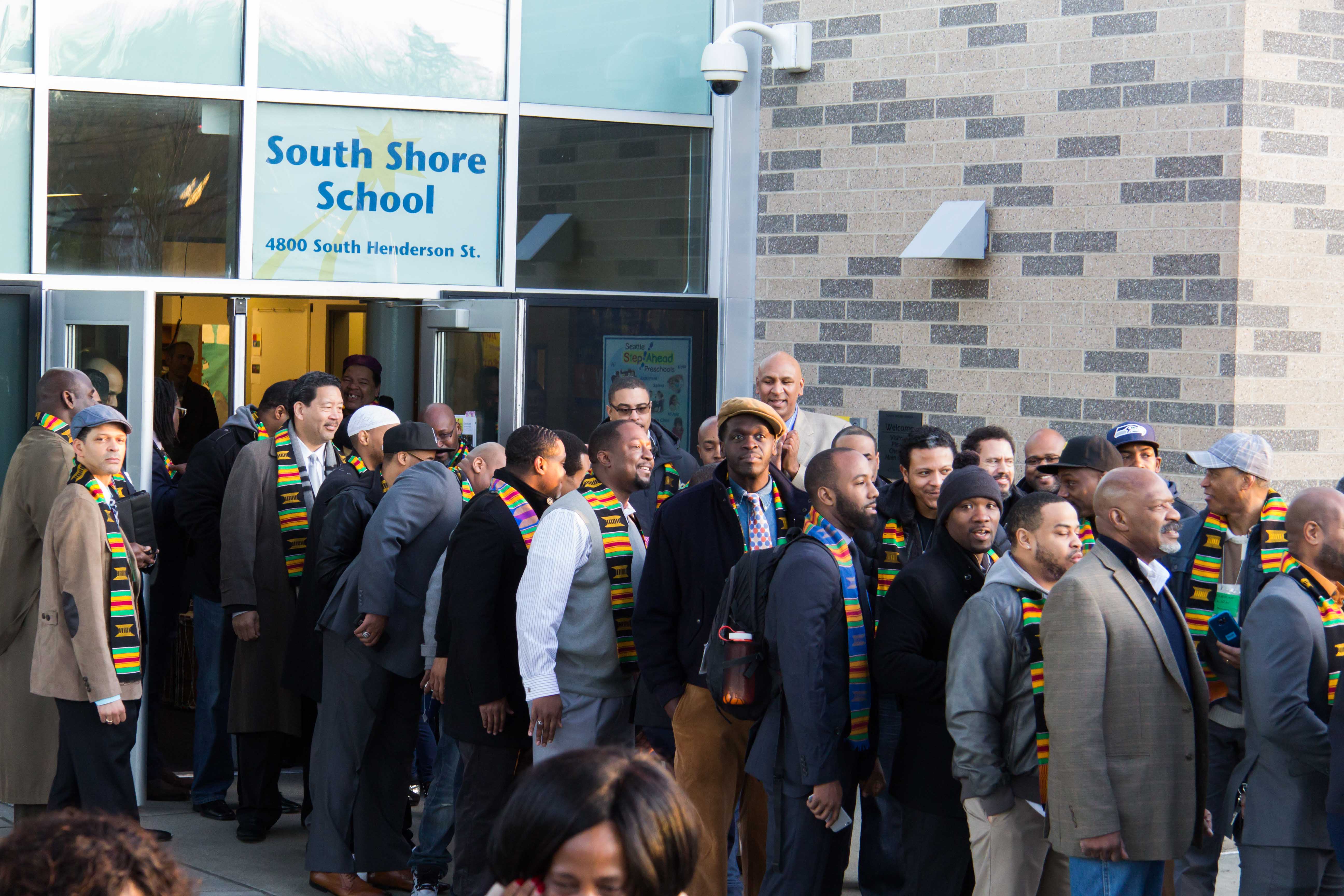 Eine Gruppe von Menschen, einige mit Mützen und Schals, steht vor dem beleuchteten Schulgebäude mit Glaswänden und Türen, versammelt zum Feiern seiner Eröffnung, wobei eine Person ein Mobiltelefon hält.