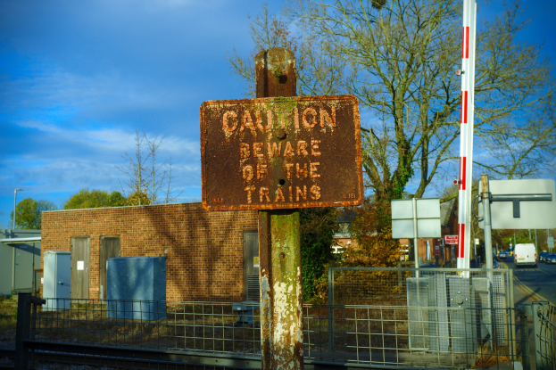 Warnschild an einem Bahnübergang-Zaun mit Bäumen, Pfählen, einem Gebäude, Containern, Fahrzeugen und einem bewölkten Himmel.