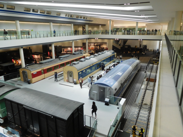 Eine große Museumsausstellung zeigt verschiedene Züge in verschiedenen Größen und Farben, mit Menschen, die vom Boden, Geländern und Säulen aus beobachten, unter Deckenlampen.