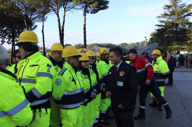 Eine Gruppe von Menschen in gelben Westen und Helmen, einige mit Mützen, steht zusammen auf einer von Bäumen umgebenen Straße unter einem klaren blauen Himmel.
