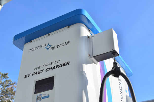 Eine Elektrofahrzeug-Ladestation mit einem blauen und weißen "V2G Fast Charger"-Schild, umgeben von Bäumen und einem Gebäude im Hintergrund unter einem klaren blauen Himmel.