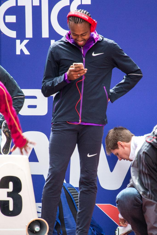 Eine Person, die mit einem Mobiltelefon in der Hand steht und lächelt, daneben eine weitere Person in Hockstellung, ein Rucksack, ein Megafon unten, eine Werbetafel im Hintergrund und eine weitere Person, die links steht.