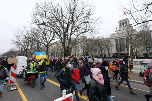 Eine große Gruppe von Menschen nimmt an einer Protestdemo auf einer Straße in Washington, D.C. teil, wobei einige Schilder und Plakate halten und andere Fahrräder fahren, sowie Schilder, Bäume und einen klaren blauen Himmel im Hintergrund.