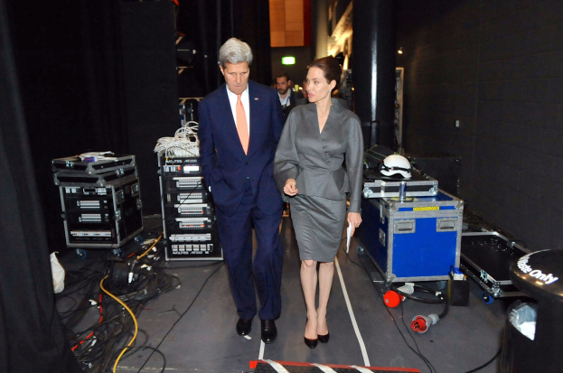 Angelina Jolie und John Kerry auf einem Boden umgeben von Menschen, Geräten, Kabeln, einem Helm auf einem Wagen, einem Mülleimer, einem Vorhang, einer Säule, einem Schild und einer Decke mit Deckenleuchten.