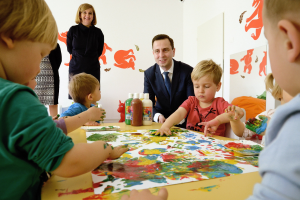 Gruppe von Kindern an einem Tisch sitzend mit Farbe an den Händen, Papieren und Flaschen auf dem Tisch, Gemälden an der Wand und einer lächelnden Frau im Hintergrund.