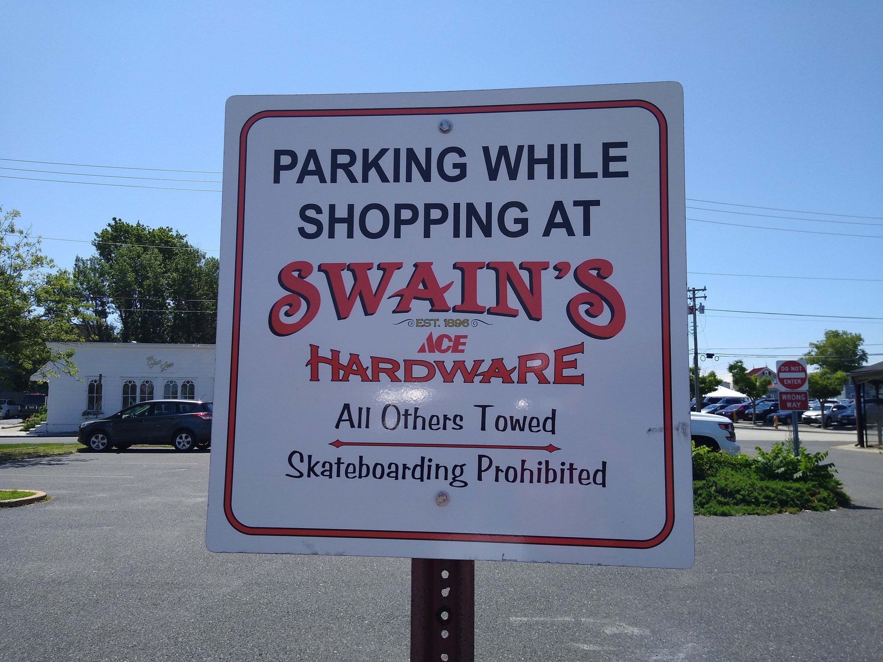 Parkplatz mit einem Schild 'Parken während des Einkaufs bei Swain's Hardware' im Vordergrund, Fahrzeuge auf der Straße, Grünfläche, Gebäude, Strommasten und ein klarer blauer Himmel im Hintergrund.