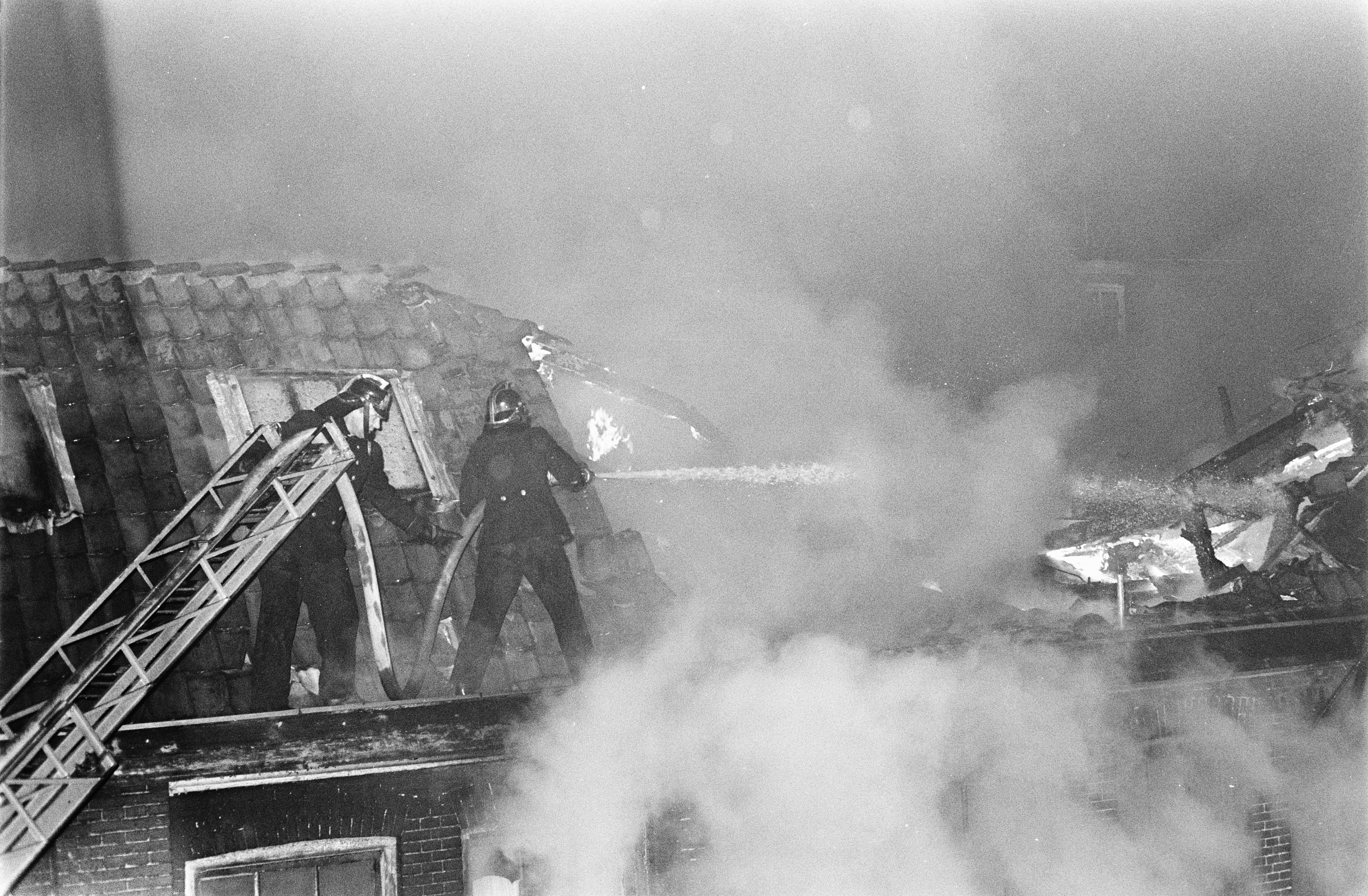 Feuerwehrleute in Helmen auf einem Gebäude, mit einer Leiter auf der linken Seite, um ein Feuer zu bekämpfen, mit Rauch, der aus dem Dach quillt.
