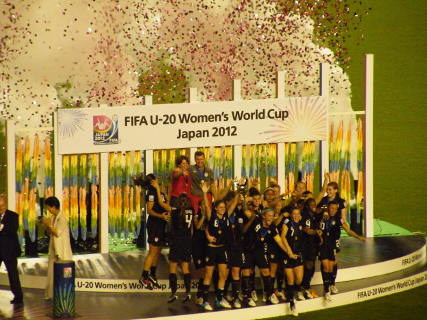 Gruppe von Frauenfußballspielerinnen in schwarzen Uniformen, die auf einer Bühne feiern, umgeben von einer Menge, mit einem dekorierten Hintergrund und grasbewachsenem Boden.