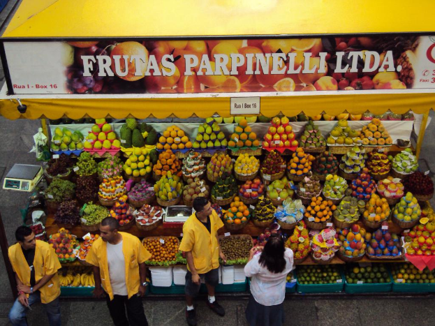 Ein Marktstand mit Körben voller Früchte, einem Banner und einer Tafel oben, einer Waage links und vier Personen, die unten auf einem Pfad stehen.