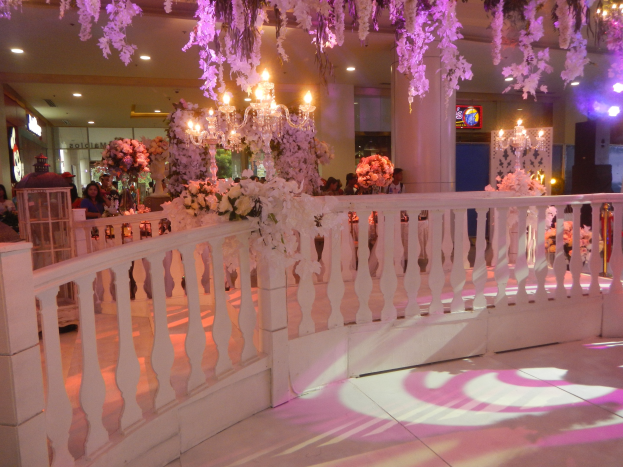 Weißes Geländer mit Blumen und Kronleuchtern geschmückt, umgeben von Gästen, Säulen und Lichter im Hintergrund sowie ein Bildschirm auf der rechten Seite des Raumes.