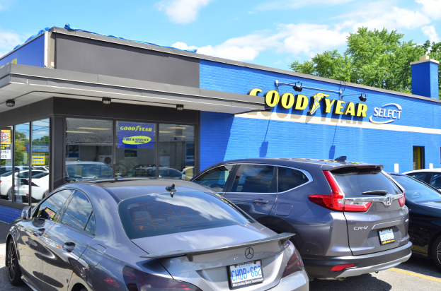 Ein Goodyear Select Autohaus mit mehreren geparkten Autos davor, das ein Gebäude mit Text und einen Hintergrund aus Bäumen unter einem bewölkten Himmel zeigt.