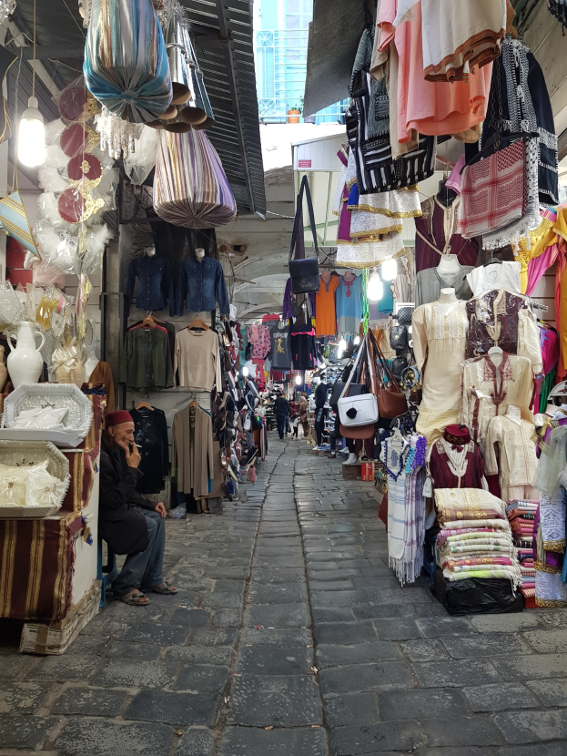 Ein belebter Straßenmarkt in der Altstadt von Jerusalem mit Ständen, die Kleidung, Taschen, Lampen und andere Gegenstände verkaufen, umgeben von Menschen, die durch die Gassen schlendern und ein Gebäude im Hintergrund.