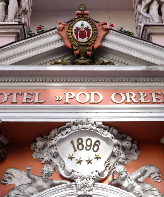 Ein Gebäude mit einer Tafel, auf der "Hotel Pod Orlen" steht, und Skulpturen an der Fassade, die detaillierte Verzierungen und ein Logo aufweisen, strahlt einen klassischen und zeitlosen Eindruck aus.