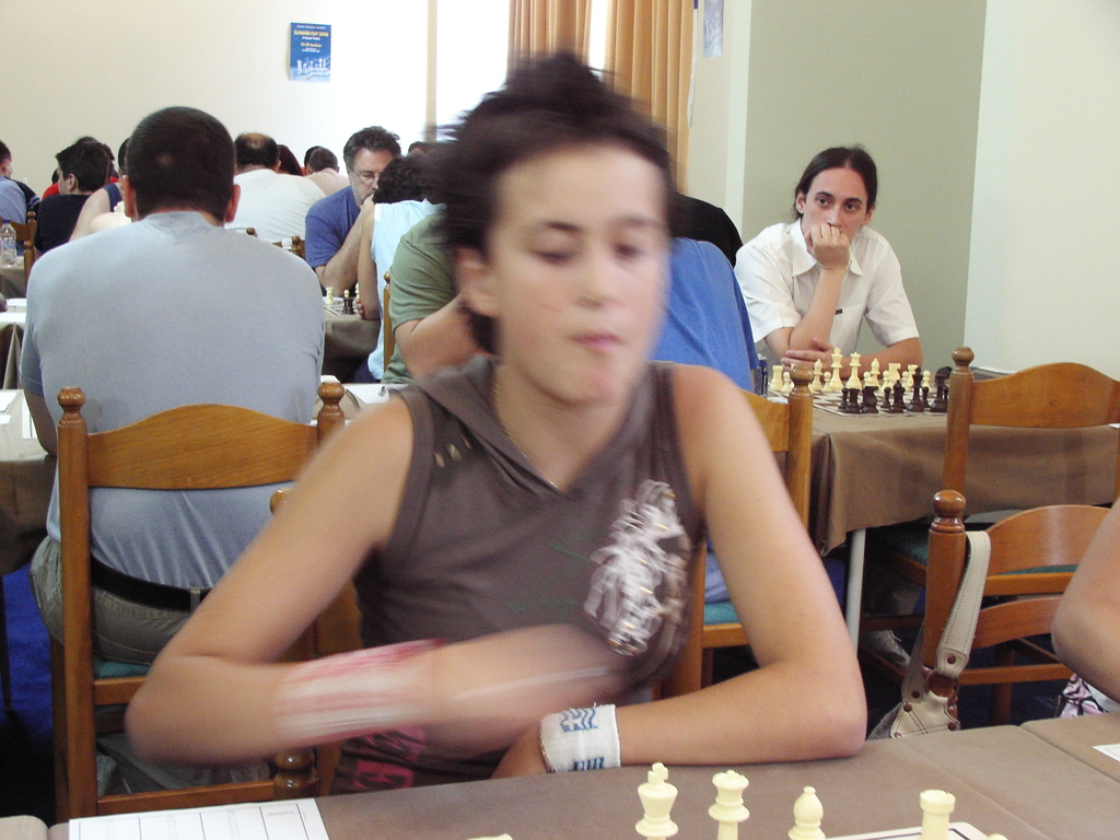 Ein Raum mit Menschen an Tischen beim Schachspielen, mit einer Vorhangwand und einem Foto in der rechten Ecke.