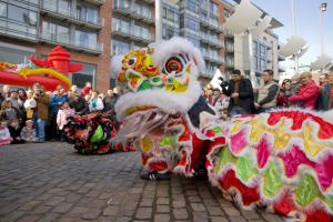 Vibrantes chinesisches Neujahrsfest in Amsterdam mit Löwen-Tanz im Vordergrund und einer Menschenmenge drumherum, Gebäuden und Laternenpfählen im Hintergrund und einem klaren blauen Himmel.