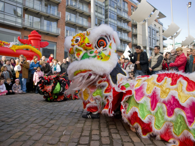 Vibrantes chinesisches Neujahrsfest in Amsterdam mit Löwen-Tanz im Vordergrund und einer Menschenmenge drumherum, Gebäuden und Laternenpfählen im Hintergrund und einem klaren blauen Himmel.