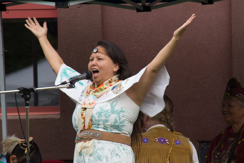 Eine Frau in einem weißen Kleid steht auf einer Bühne, spricht in ein Mikrofon, mit einer Wand und einem Eisenrahmen dahinter.