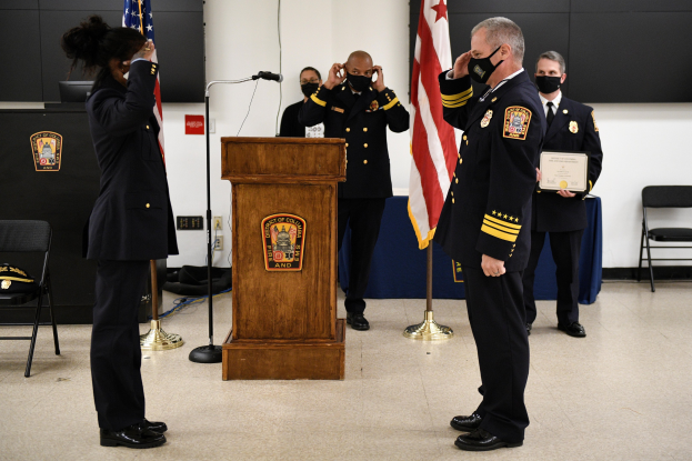 Gruppe uniformierter Personen in Masken vor einem Rednerpult mit Mikrofon während einer Vereidigung eines neuen Feuerwehrchefs, der ein Zertifikat hält, umgeben von Fahnen, Stühlen, Tischen und einer Wand.