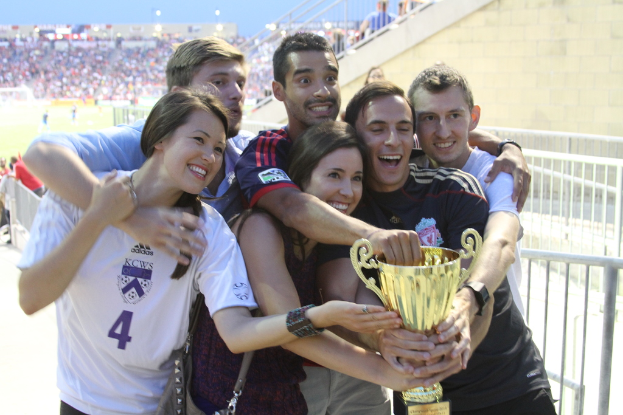 Sechs Personen, zwei Frauen und vier Männer, halten einen Pokal und lächeln, posieren für ein Foto vor einem Eisengeländer, mit einer Wand und einem Geländer oben rechts, Menschen und Lichter im Hintergrund, möglicherweise in einem Stadion.