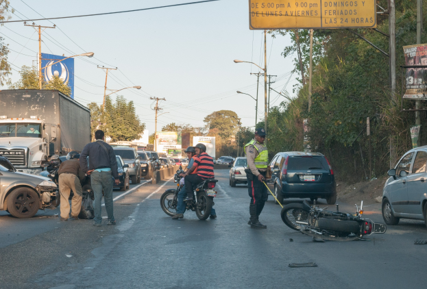 Eine Gruppe von Menschen steht in der Nähe eines verunglückten Motorrads auf der Fahrbahn mit mehreren Fahrzeugen, darunter ein Lastwagen, im Hintergrund, umgeben von Bäumen, Masten, Lichtern, Schildern und dem Himmel.