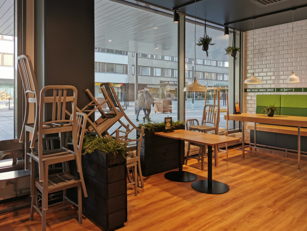 Ein Restaurantinnenraum mit hölzernen Tischen und Stühlen, großen Fenstern, Pflanzen und Beleuchtung, mit Blick auf eine Berliner Straße mit Fußgängern, Gebäuden und Strommasten.