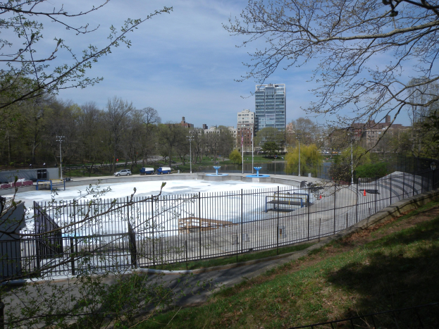 Eisstadion mit Metallzaun in einem Park mit Bäumen, Gras, Fahrzeugen, Straßenlaternen, einem Gewässer, Gebäuden und einem bewölkten Himmel.