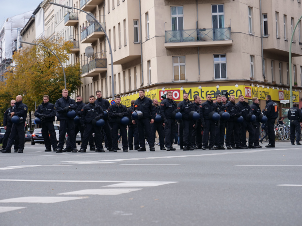 Eine Gruppe von Polizisten in schwarzen Uniformen mit blauen Helmen steht auf einer Straße, die von Laternenmasten, Bäumen und Glasfenstergebäuden gesäumt ist, unter einem klaren blauen Himmel.