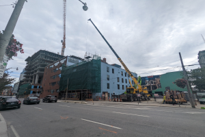 Eine belebte Stadtstraße mit Fahrzeugen, Fußgängern, Straßeninfrastruktur und Gebäuden, mit einem Baukran, der im Hintergrund eine Struktur anhebt.