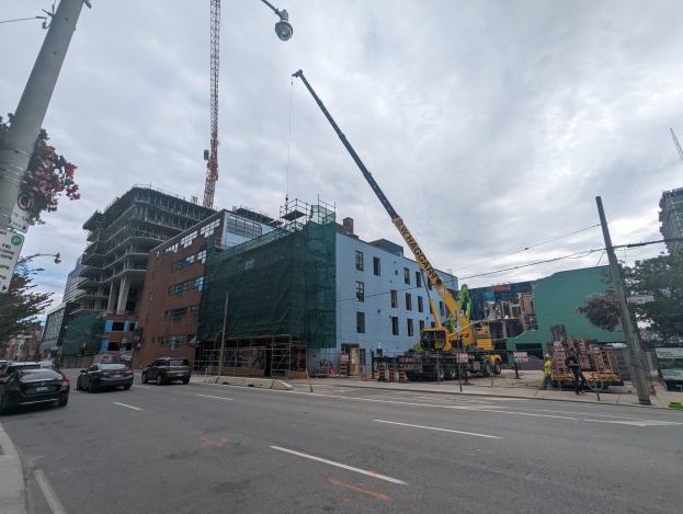 Eine belebte Stadtstraße mit Fahrzeugen, Fußgängern, Straßeninfrastruktur und Gebäuden, mit einem Baukran, der im Hintergrund eine Struktur anhebt.