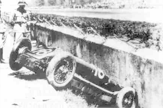 Ein Schwarz-Weiß-Foto eines verunglückten Rennwagens mit zwei Personen auf der linken Seite und Bäumen im Hintergrund.