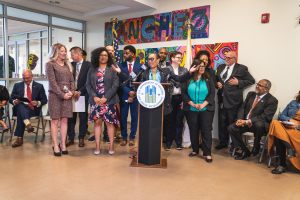Gruppe von Menschen vor einem Podium mit einem Mikrofon und einer Tafel mit der Aufschrift "Sancho City Councilman Joins City Council", mit sitzenden und stehenden Teilnehmern, Flaggen, Feuerlöscher und Deckenbeleuchtung.