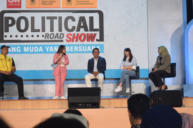 Eine Gruppe von Menschen sitzt auf einer Bühne mit einer Frau in der Mitte, die ein Mikrofon hält, einer Zuschauermenge im Vordergrund und einem Banner mit der Aufschrift "Politischer Roadshow 2019" im Hintergrund.