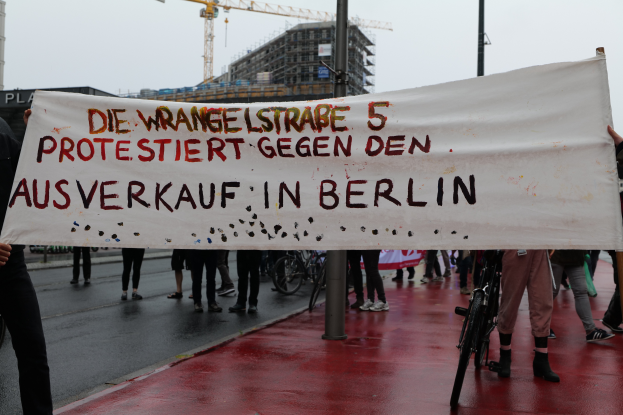 Eine Gruppe von Menschen steht auf einer Straße und hölt ein Banner mit der Aufschrift "Die Wrangellstraße 5 protestiert gegen den Ausverkauf in Berlin". Im Hintergrund sind Fahrráder, Stäbe, Gebäude und ein Kran zu sehen, öber ihnen der Himmel.