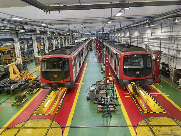 Zwei Züge auf den Schienen in einer großen Industrieanlage mit Maschinen und Geräten, mit einer Wand mit Rohren und Deckenbeleuchtung auf der rechten Seite.