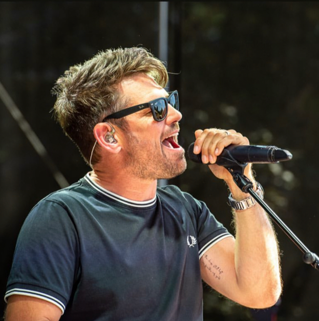 Ein Mann in schwarzem T-Shirt und Sonnenbrille singt in ein Mikrofon mit Ständer vor sich, mit einem unscharfen, träumerischen Hintergrund.