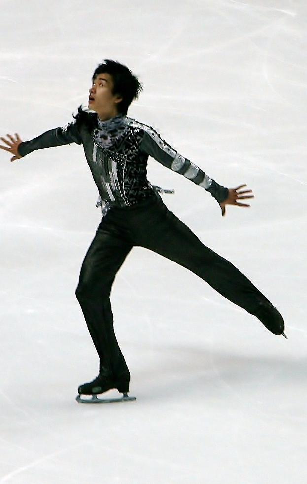 Mann in schwarzer Eiskunstlaufbekleidung gleitet über eine Eisfläche und führt eine Choreografie aus.